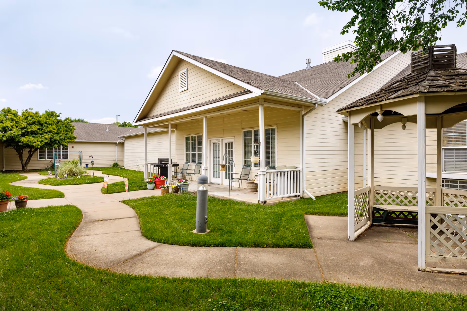 Outdoor view of a senior living facility with beige buildings, a covered porch with chairs, a gazebo, paved walkways, green grass, and potted plants under a partly cloudy sky.