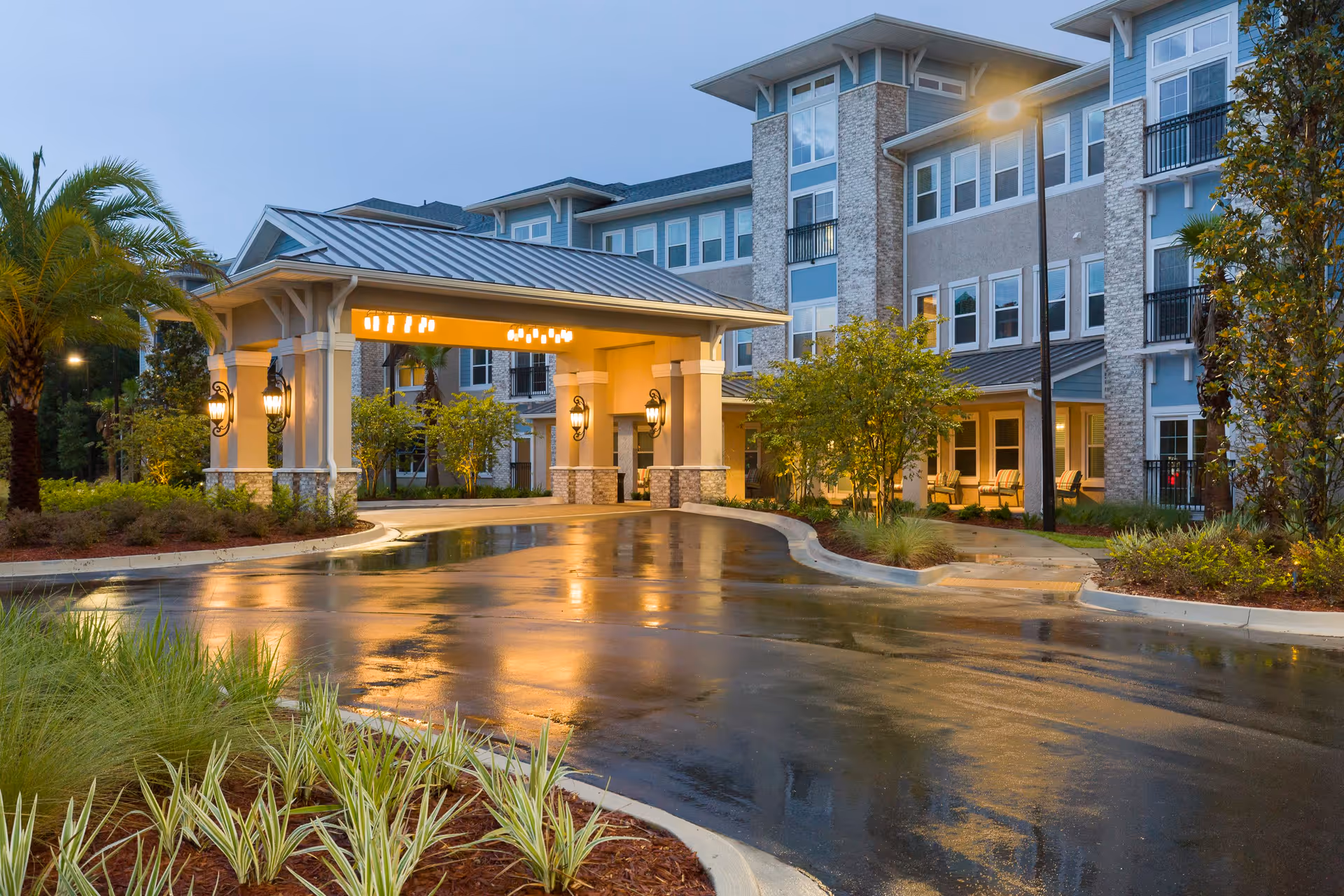 Entrance of a senior living facility named HarborChase of Mandarin during dusk, showing a covered drop-off area with warm lighting, surrounded by landscaped greenery and a multi-story building in the background.