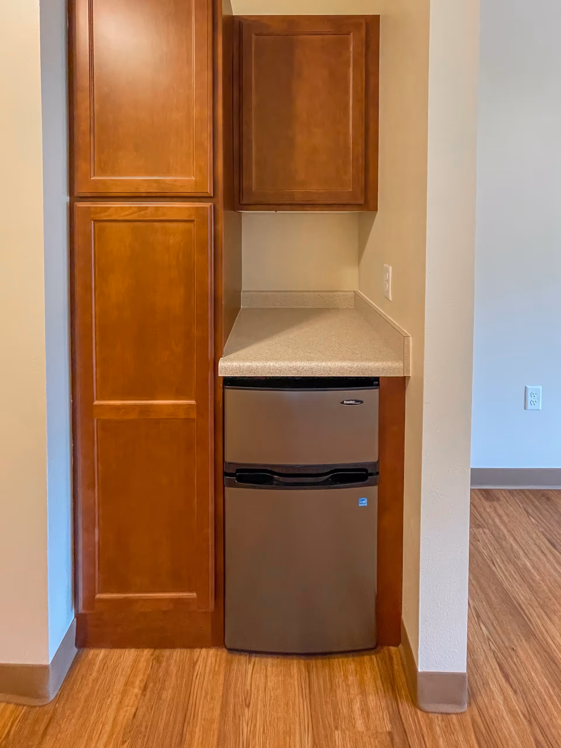 A small kitchen area with wooden cabinets, a beige countertop, and a compact stainless steel refrigerator underneath the counter. The floor is wood, and there is a light-colored wall with an electrical outlet visible.