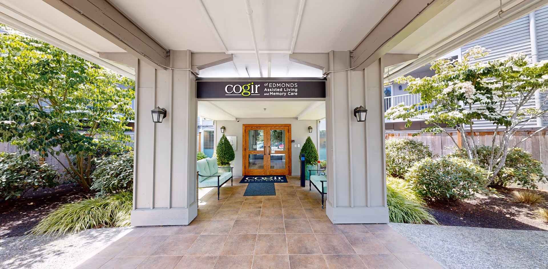Covered entrance area of Aegis of Edmonds Assisted Living facility with tiled floor, two green cushioned benches on either side, potted plants, and double wooden doors at the end. The entrance is flanked by greenery and trees on both sides.