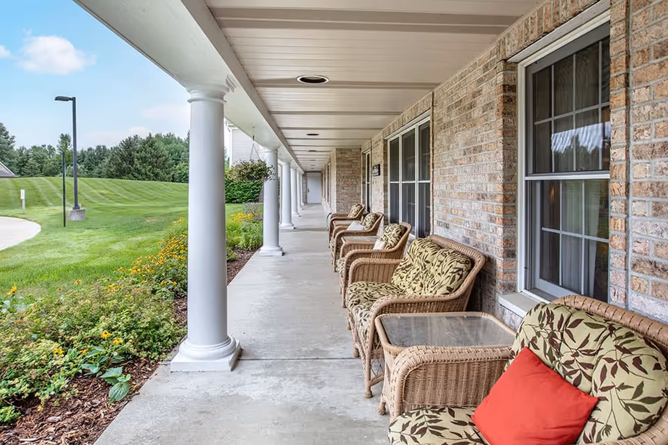 Covered outdoor walkway with white columns and wicker chairs with cushions lined up against a brick wall, overlooking a green lawn and garden with yellow flowers under a partly cloudy sky.