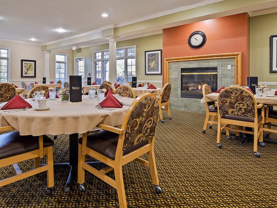 A dining room in an assisted living facility with round tables covered in beige tablecloths, each set with glasses, cups, and red folded napkins. The chairs have wooden frames and patterned upholstery. There is a fireplace with a clock above it on an orange accent wall, and large windows letting in natural light.