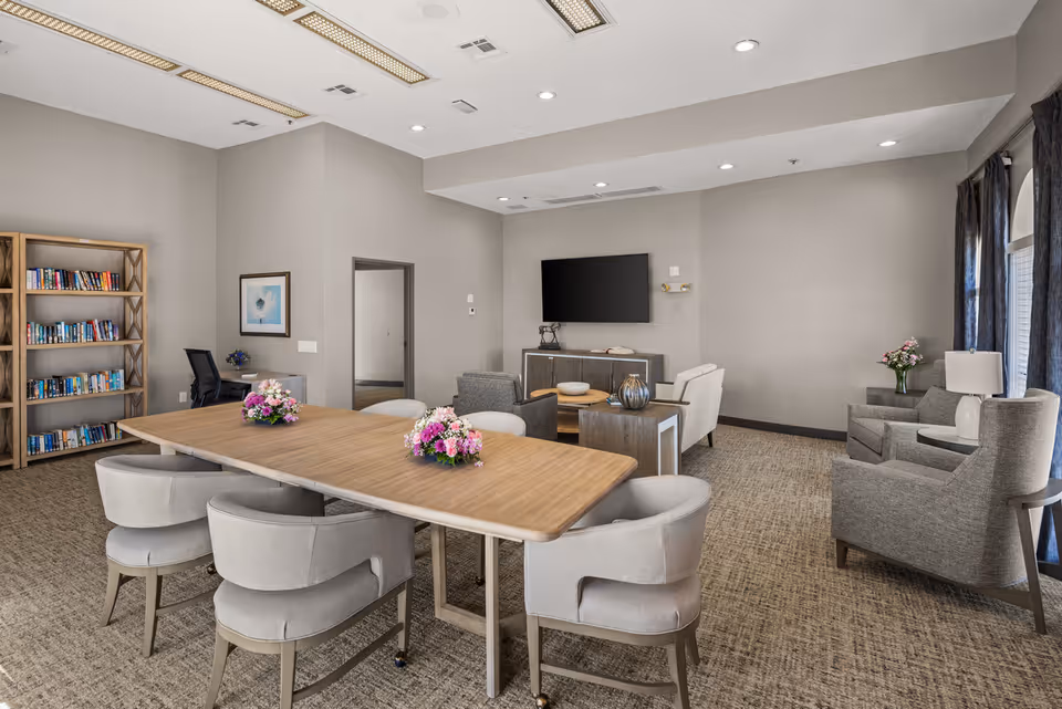 A spacious senior living community room featuring a large wooden table with six upholstered chairs, decorated with three floral centerpieces. In the background, there is a seating area with armchairs, a coffee table, a TV mounted on the wall, and a bookshelf filled with books. The room has neutral-colored walls, carpeted floor, and large windows with dark curtains allowing natural light.