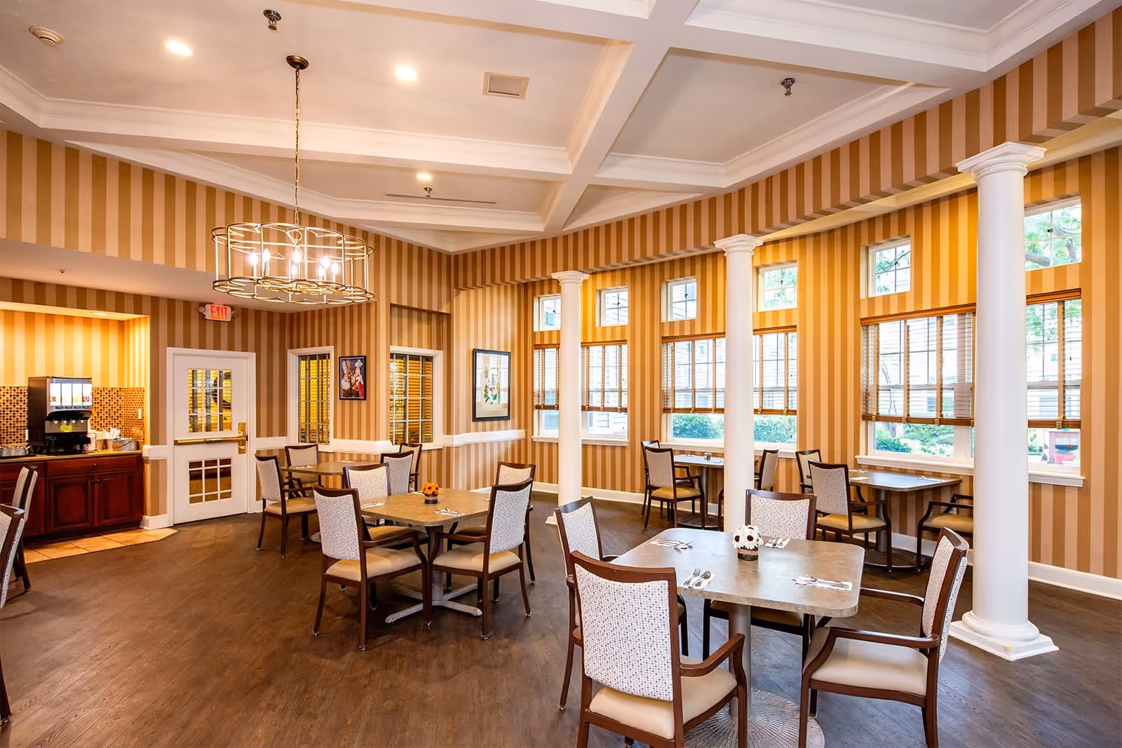 Dining room with several tables and chairs, striped wallpaper, tall white columns, and large windows.