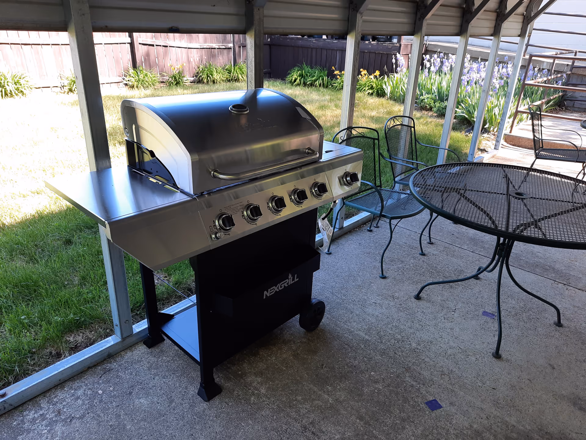 Stainless steel gas grill on a covered patio beside a round metal table and chairs with a grassy yard and flowers beyond.