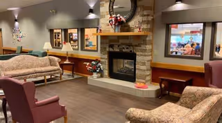 A cozy senior living facility common area with multiple upholstered chairs and sofas arranged around a stone fireplace. The fireplace has a wooden mantle decorated with flowers and a round mirror above it. The room features warm lighting, wood flooring, and windows looking into another room where people are seated.