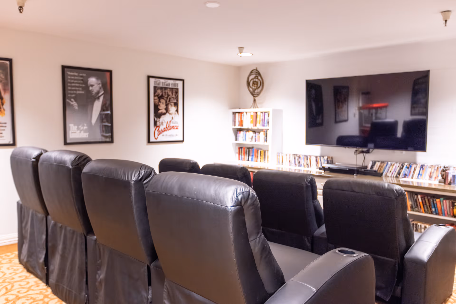 A cozy media room with two rows of black leather recliner chairs facing a large flat-screen TV mounted on the wall. The room has movie posters on the walls and shelves filled with DVDs and books underneath the TV and in a corner bookshelf.