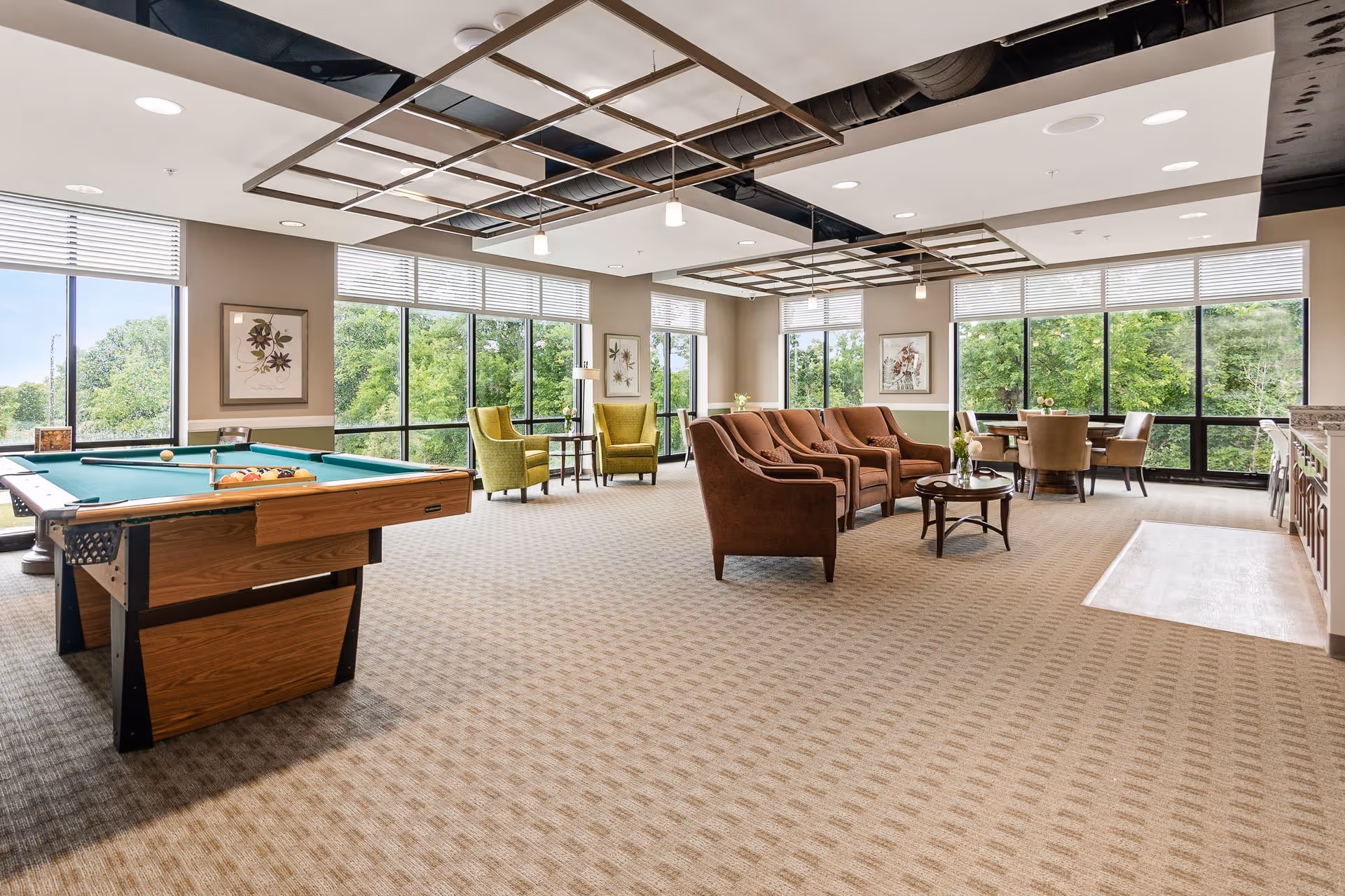 A spacious and well-lit common area in a senior living facility featuring a pool table, several armchairs, a small round table with chairs, large windows with views of green trees, and modern ceiling light fixtures.