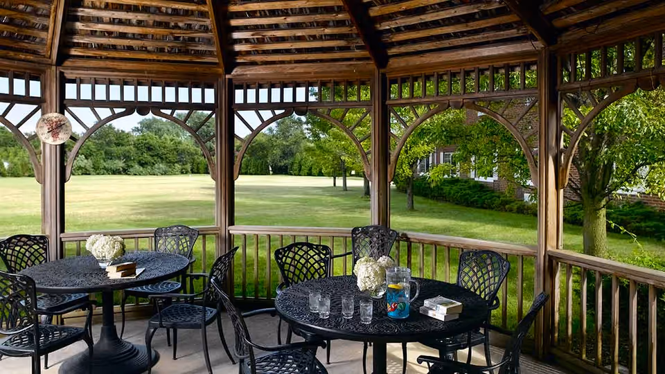 A wooden gazebo with a slatted roof and decorative arches, containing two black metal round tables each surrounded by black metal chairs. On the tables are glass pitchers with water and lemon slices, drinking glasses, white floral centerpieces, and books. The gazebo overlooks a large grassy lawn with trees and a brick building partially visible in the background.