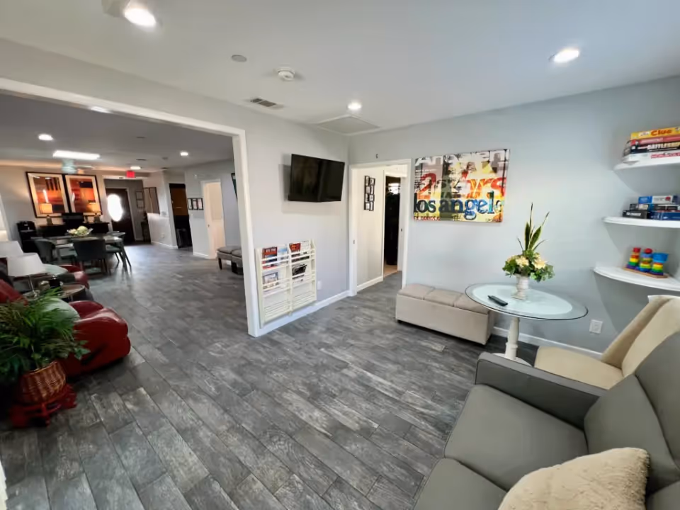 Interior view of a senior living facility lounge area with gray tiled flooring, light gray walls, and recessed ceiling lights. The room features a wall-mounted TV, a glass-top round table with a flower arrangement, cushioned chairs, a small bench, and wall-mounted shelves holding board games. In the background, there is a dining area with a table and chairs, artwork on the walls, and a potted plant near a red recliner chair.