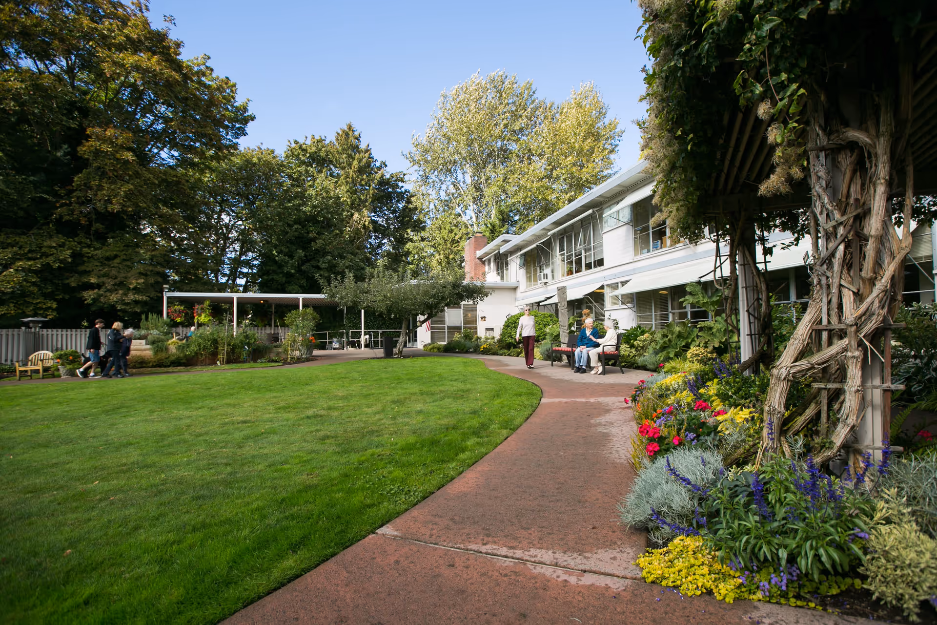 Outdoor garden area at Ida Culver House Ravenna with a curved paved walkway, green lawn, colorful flower beds, and a white two-story building in the background. Several people are walking and sitting on benches enjoying the sunny day.