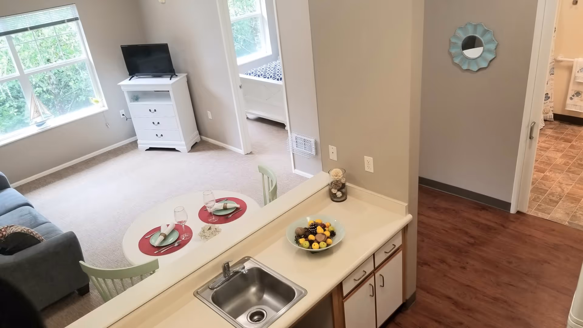 View of a senior living facility apartment showing a small kitchen area with a sink and a bowl of fruit on the counter. Beyond the kitchen is a living room with a round table set for two, a couch, a window with natural light, and a TV on a white dresser. A bedroom with a bed is visible through an open doorway, and a hallway leads to a bathroom with tiled flooring.