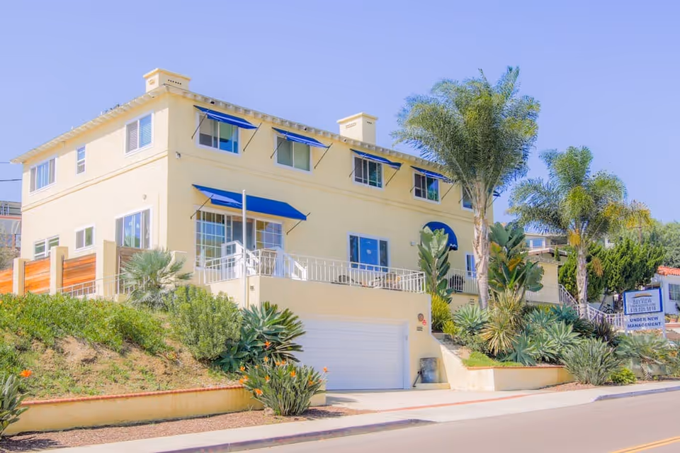 Exterior view of a two-story yellow building with blue awnings over the windows and doors, surrounded by palm trees and various green plants. There is a garage door at the lower level and a sign near the street that reads 'BayView Senior Assisted Living Under New Management.'