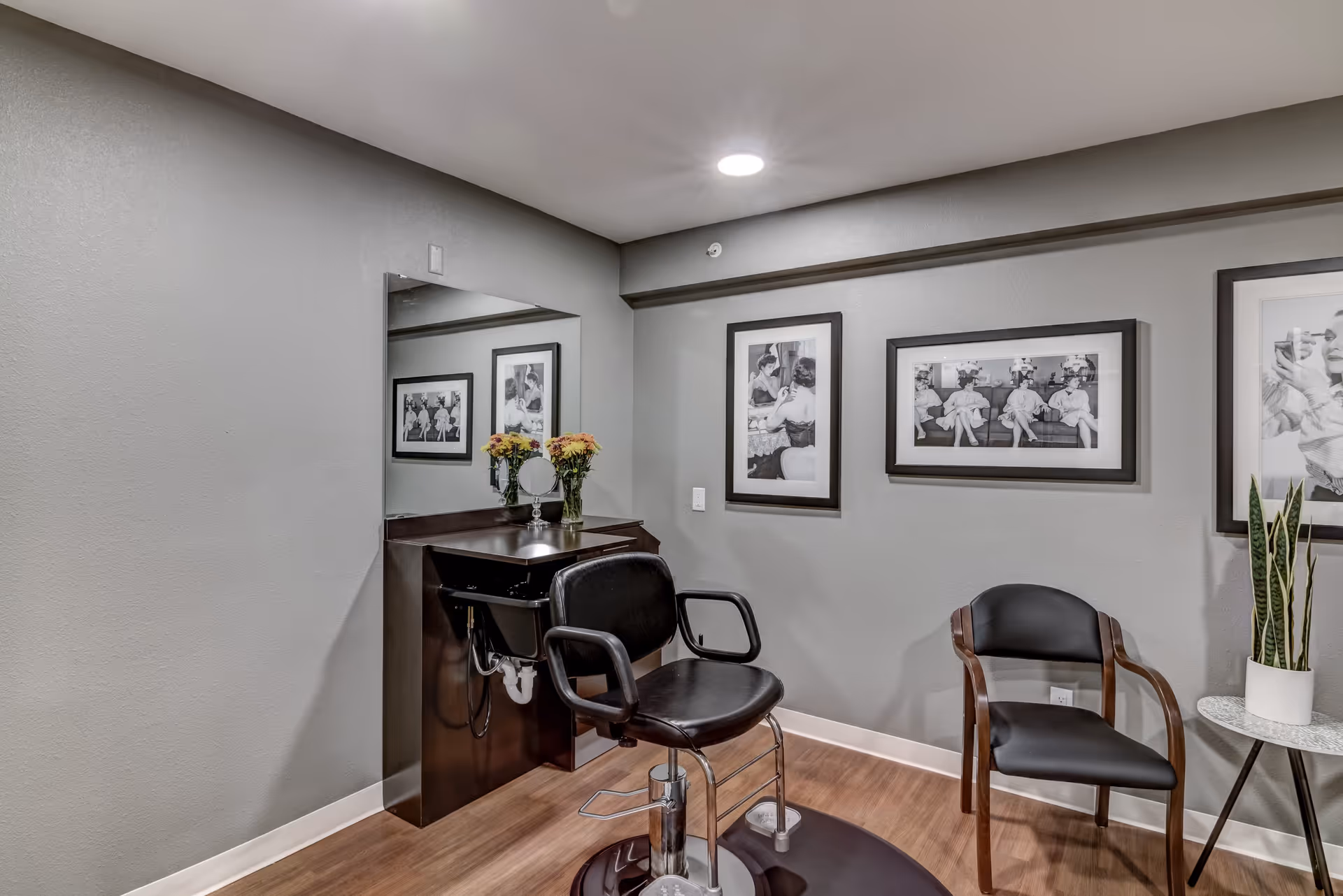A small salon area with a black salon chair in front of a mirror and a sink. There is a vase with flowers on the counter, a black chair with wooden armrests, a small round table with a potted plant, and three black and white framed photos on the gray walls.