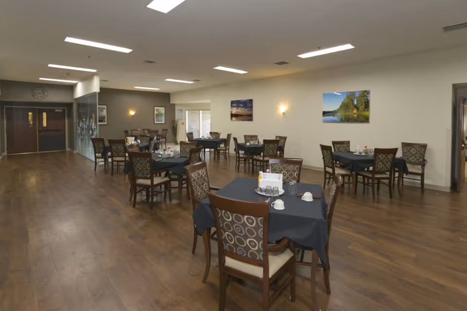 Spacious dining room with multiple tables set with dark tablecloths and patterned chairs on wood flooring.