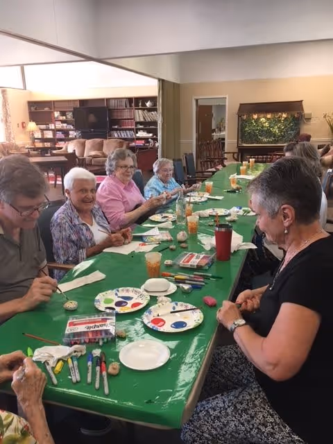 A group of elderly people sitting around a long table covered with a green tablecloth, engaging in a craft activity with paint markers and small objects. The room has bookshelves, a television, and a decorative wall feature in the background.
