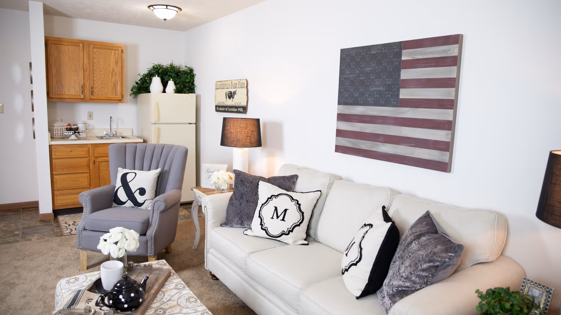 A cozy living area featuring a beige sofa with decorative pillows, a gray armchair with a pillow, a small wooden side table with a lamp, and a coffee table with a teapot and cup. In the background, there is a small kitchenette with wooden cabinets, a sink, and a refrigerator. A rustic American flag wall art hangs above the sofa, and there are plants and decorative items placed around the room.