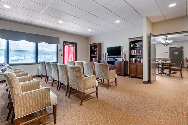A spacious living room area with two rows of beige patterned armchairs facing a large flat-screen TV mounted on a wooden cabinet. The room has a carpeted floor, a window with blinds and a valance, and built-in wooden shelves flanking the TV. An adjacent dining area with tables and chairs is visible through an open doorway.