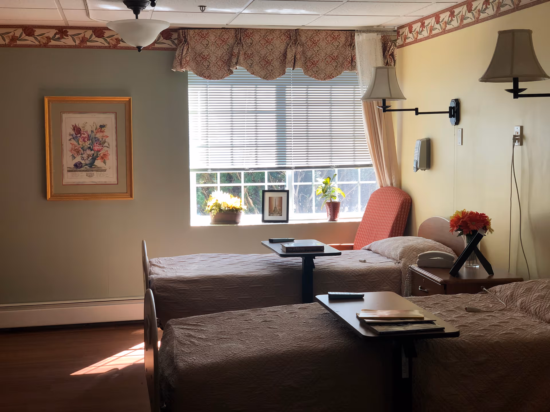A room with two single beds covered in beige bedspreads, each with an overbed table holding books and a remote control. There is a window with blinds and floral valance letting in natural light. A red upholstered chair is positioned near the window. The walls are decorated with a floral border near the ceiling, a framed floral painting, and two wall-mounted lamps with beige shades. A nightstand between the beds holds a telephone and a vase with flowers.