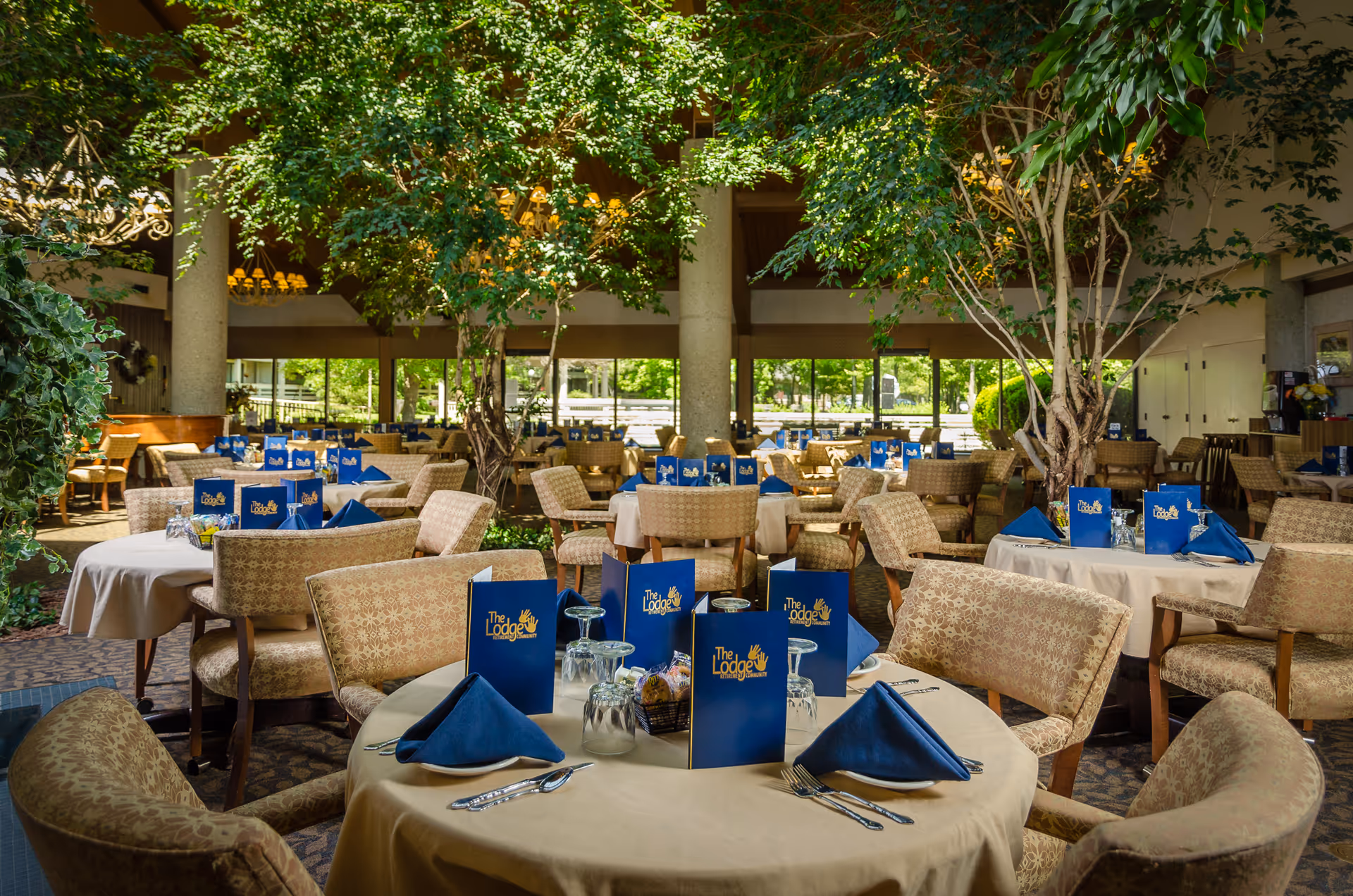 A spacious dining room at The Lodge Retirement Community with round tables covered in beige tablecloths, each set with blue napkins, silverware, upside-down glasses, and blue menus featuring the facility's logo. The room has large windows letting in natural light and several indoor trees adding greenery to the space.