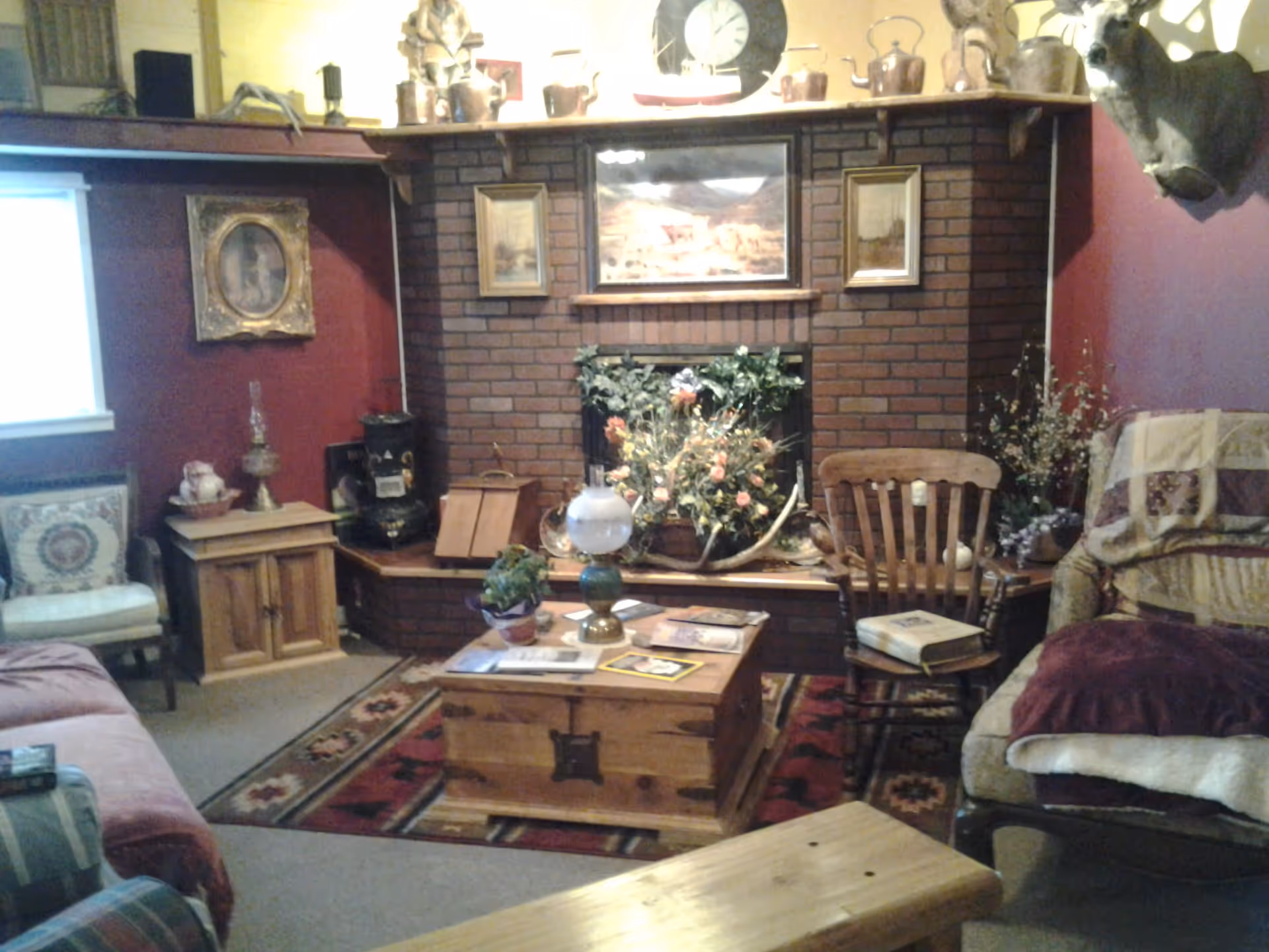 Cozy living room with a brick fireplace decorated with framed pictures and various antique items on the mantel. There is a wooden chest serving as a coffee table with books and a lamp on it, surrounded by a wooden rocking chair, cushioned armchairs, and a patterned rug. The walls are painted a deep red color and there are floral arrangements and decorative pillows adding to the warm atmosphere.