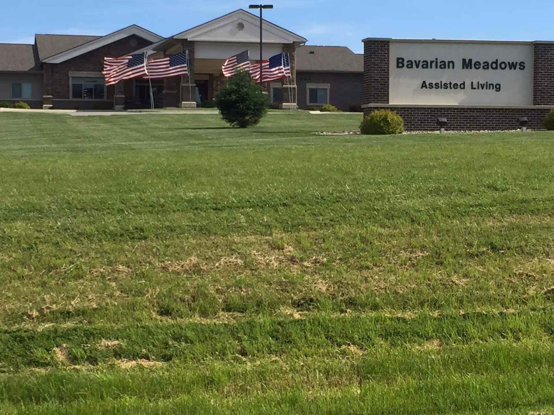 Exterior view of Bavarian Meadows Assisted Living facility with a large green lawn in the foreground, a sign displaying the facility's name, and several American flags near the entrance of the building.