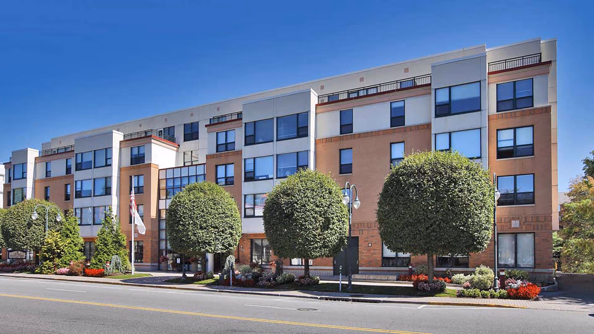 Exterior view of a multi-story senior living facility building with large windows, neatly trimmed round trees, landscaped flower beds, and a clear blue sky.