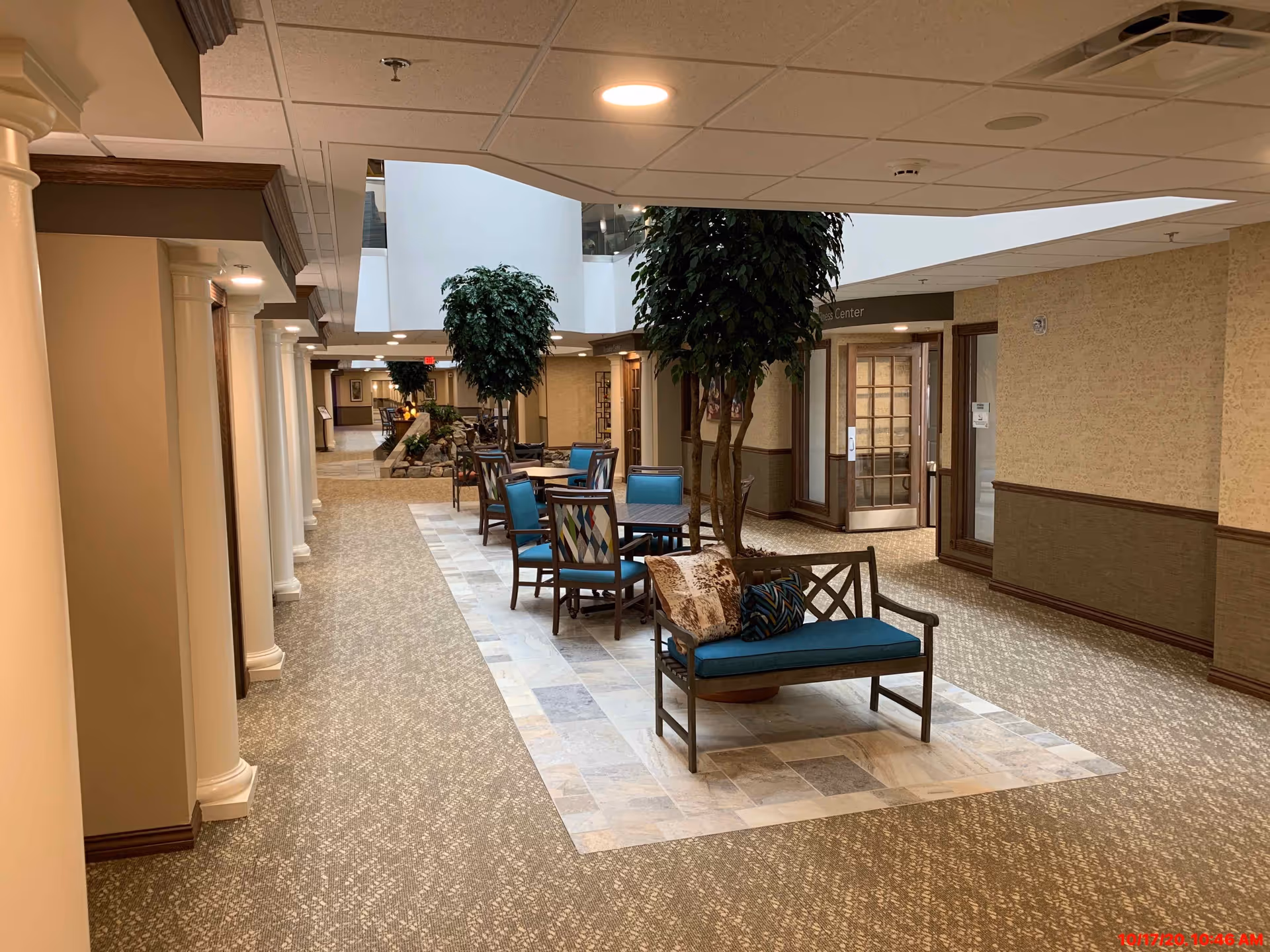 Interior hallway of a senior living facility with a seating area featuring blue cushioned chairs and a bench with pillows around indoor trees. The hallway has beige walls, carpeted floors with a tiled section under the seating area, and white columns along the left side. There is a glass door labeled 'Center' on the right side.