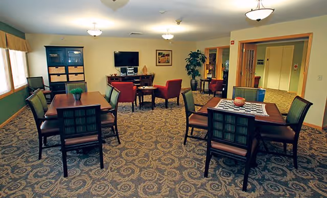 Well-lit community room with multiple tables and chairs, a seating area with armchairs and a TV, and decorative plants.