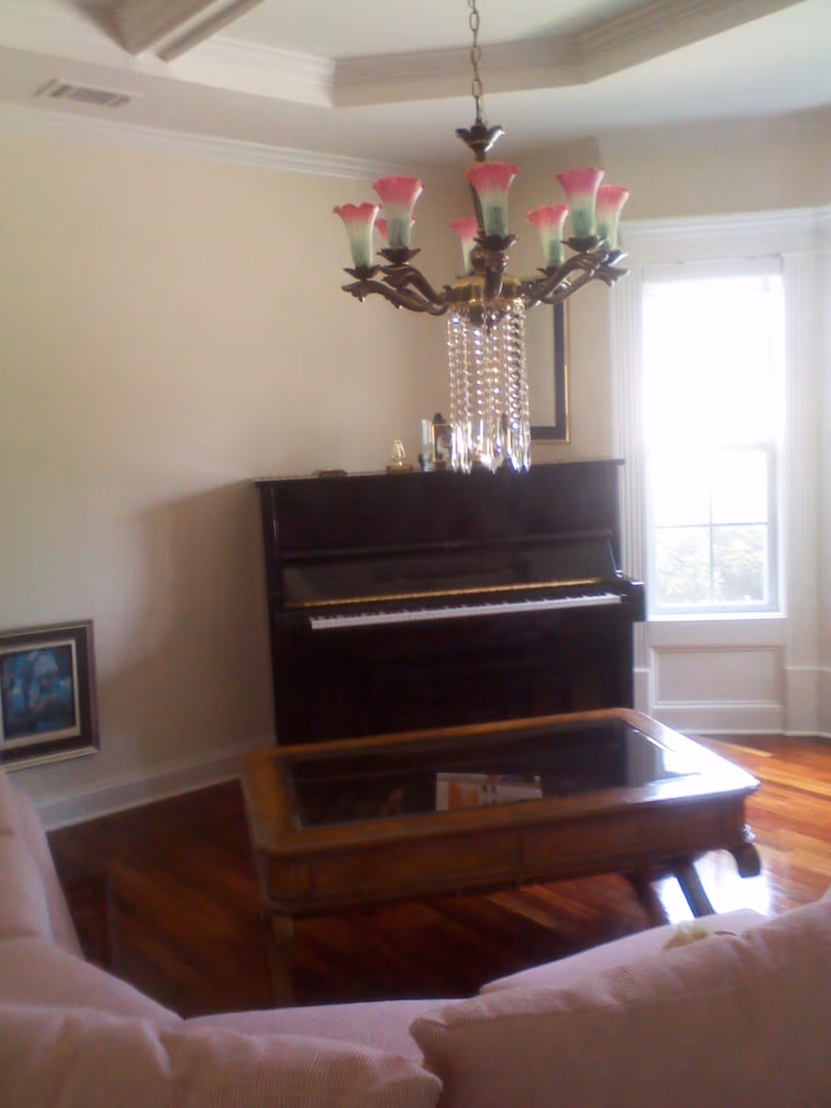 A living room with a dark upright piano against the wall, a wooden coffee table with a glass top in front of a light-colored sofa, a window with white trim letting in natural light, and a chandelier with pink and green floral-shaped lights hanging from the ceiling.