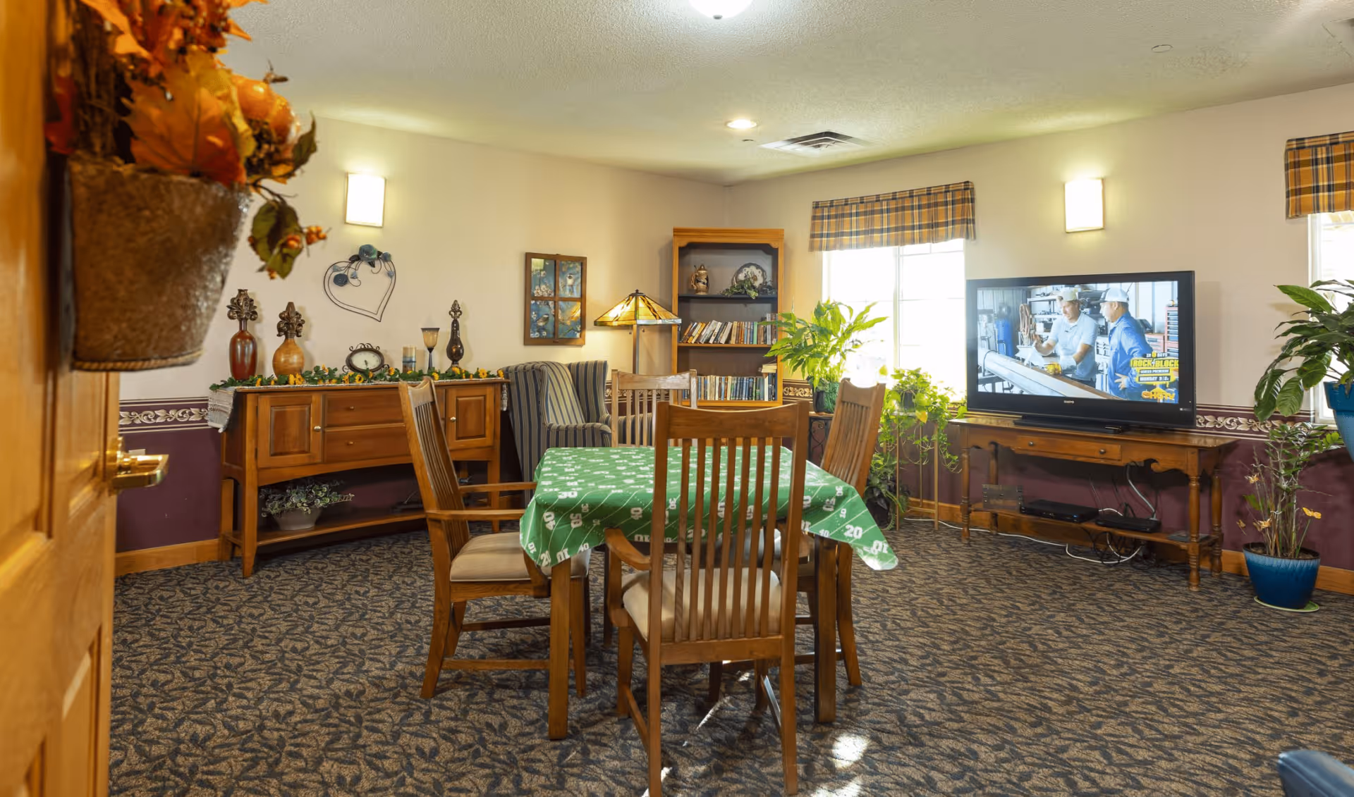 A cozy common room in an assisted living facility featuring a wooden table with four chairs covered by a green tablecloth, a TV on a wooden stand showing a program with two men, a bookshelf filled with books, a striped armchair, and various plants and decorative items around the room.