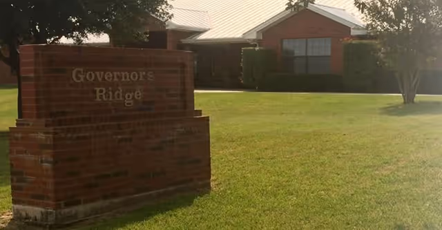 A brick sign reading 'Governor's Ridge' in front of a single-story brick building with a metal roof, surrounded by a well-maintained grassy lawn and trees.