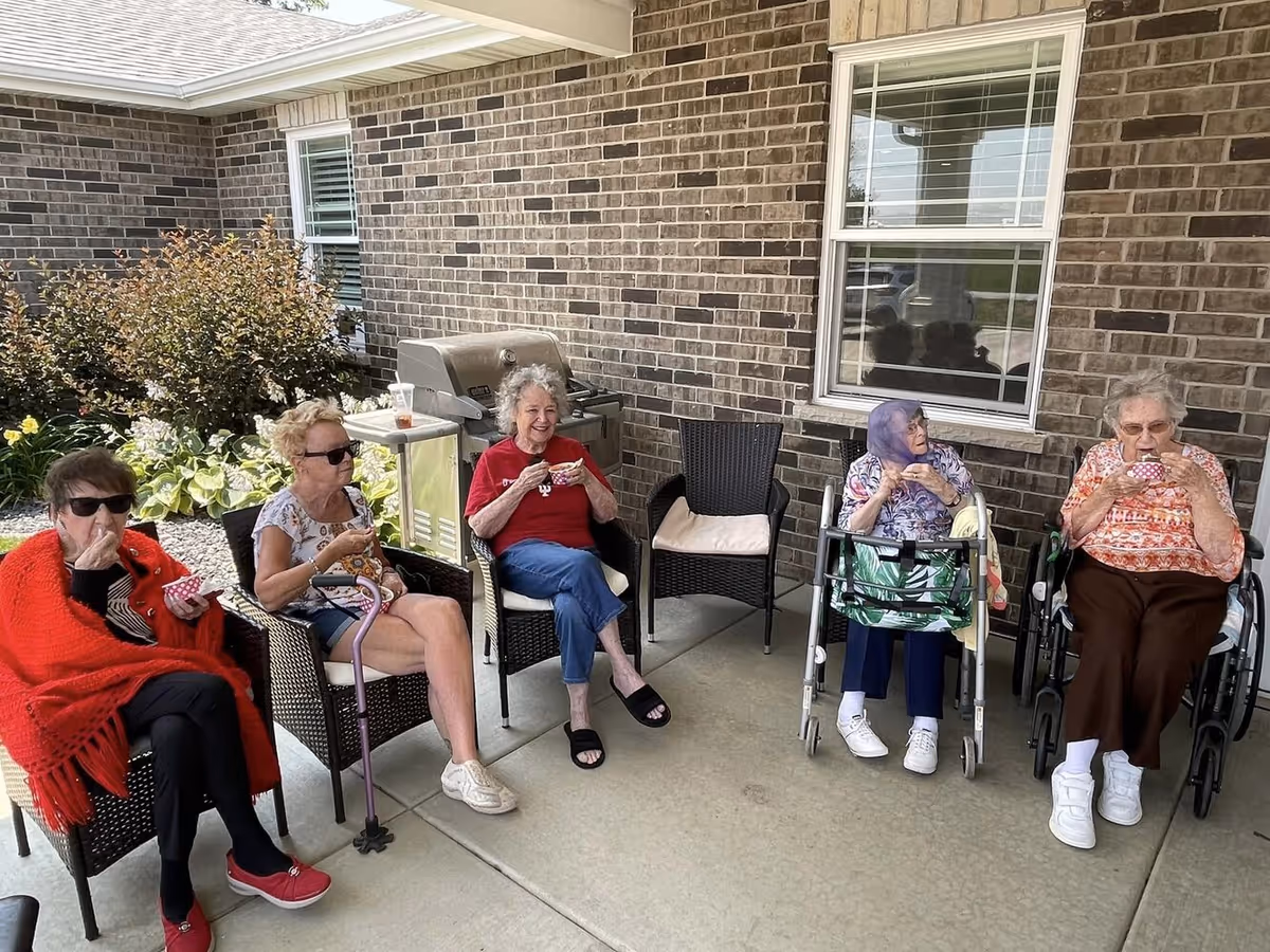 Five elderly women sitting outside on a covered patio against a brick wall, enjoying snacks. Two women are using walkers, and one woman is wrapped in a red shawl. There is a grill and some plants in the background.