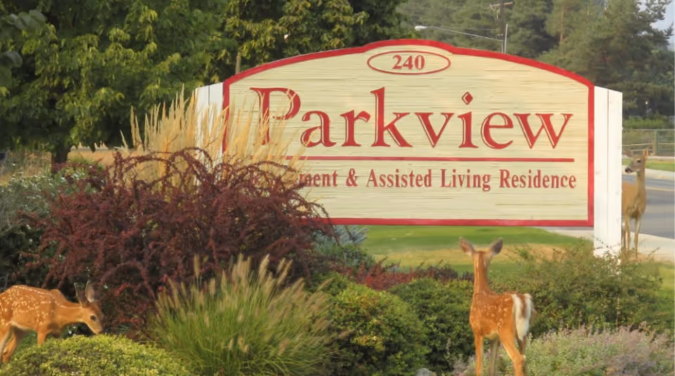 A large wooden sign for Parkview Independent & Assisted Living Residence surrounded by bushes and grass, with three deer visible near the sign and a road in the background.
