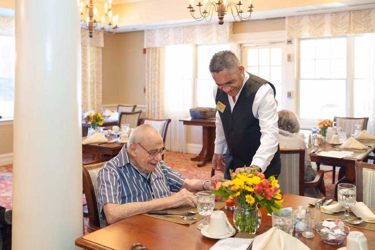 An elderly man sitting at a dining table in a well-lit dining room with a smiling staff member standing beside him. The table is set with glasses, napkins, cutlery, and a vase of colorful flowers. Other tables and chairs are visible in the background with similar settings.