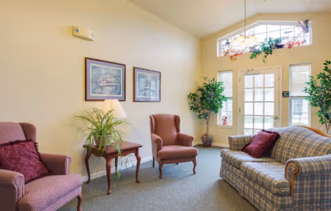 A cozy living room area with two upholstered armchairs and a plaid sofa with red cushions. There is a wooden side table with a lamp and a potted plant on it. Two framed pictures hang on the beige wall. The room has a large window above a glass door letting in natural light, with two tall potted plants on either side of the door.