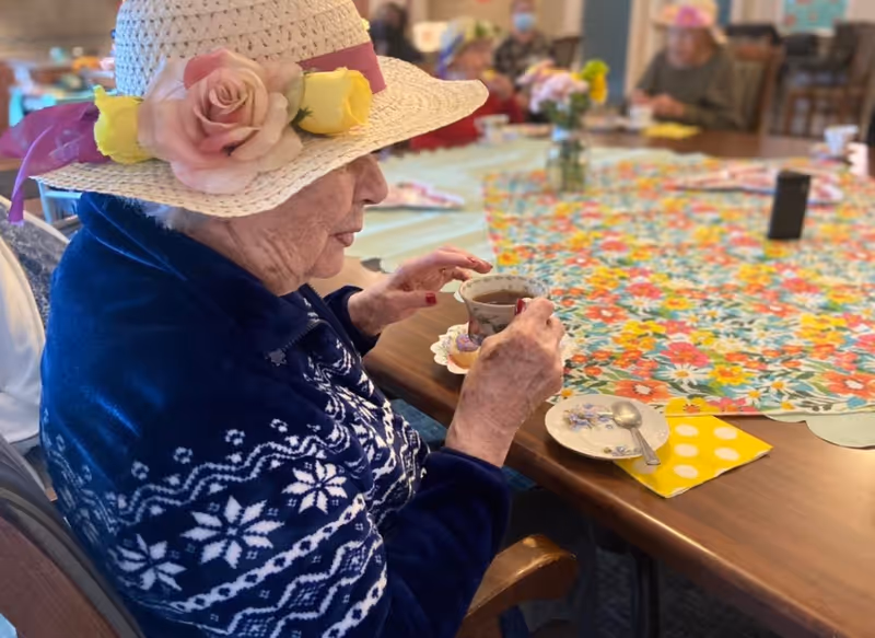 An elderly woman wearing a white hat decorated with pink and yellow flowers and a blue sweater with white snowflake patterns is sitting at a table holding a teacup. The table is covered with a colorful floral tablecloth and has a small plate with a spoon and a yellow polka dot napkin. Other elderly people are seated around the table in the background.