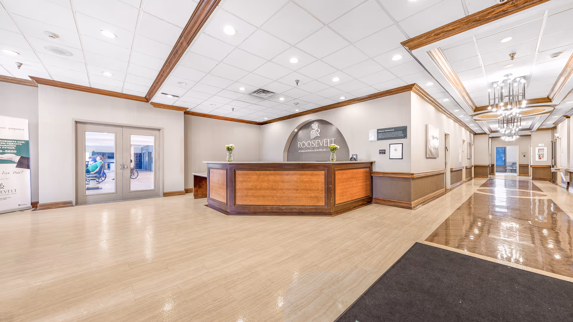 Reception area of Roosevelt Rehabilitation & Healthcare Center with a wooden front desk, light-colored flooring, and a hallway extending to the right. The center's logo and name are displayed on the wall behind the desk. Double glass doors are visible to the left, leading to another room where a person in a wheelchair can be seen.