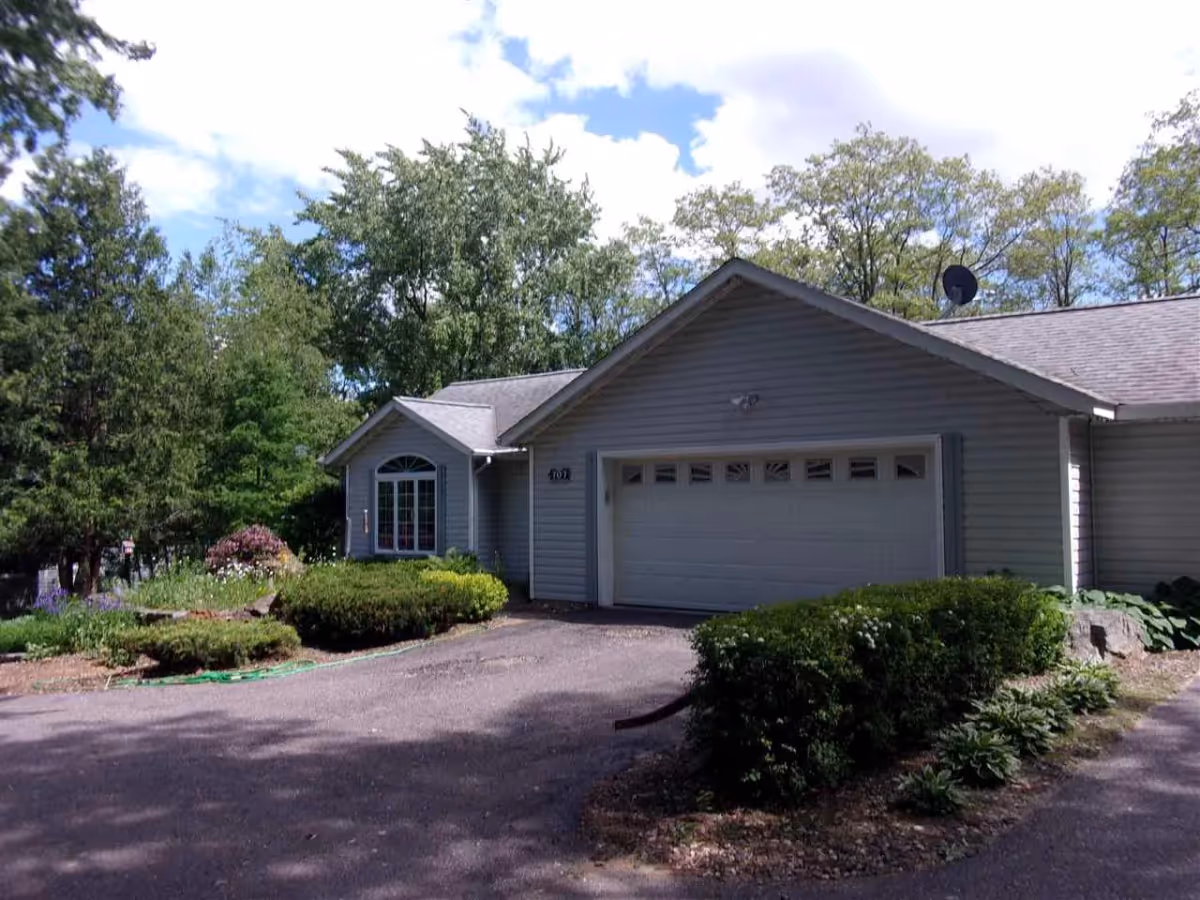Exterior view of a single-story residential building with gray siding, a double garage door, and a driveway. The building is surrounded by green bushes, trees, and a partly cloudy sky.