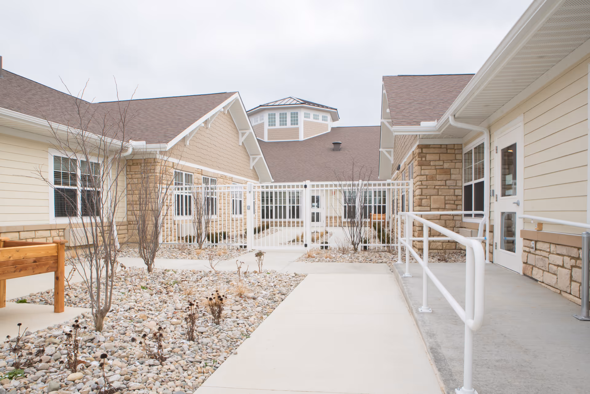 Outdoor courtyard area of a senior living facility with beige buildings featuring stone accents and white trim. The courtyard has a concrete walkway, a white metal fence gate, leafless small trees, and a wooden planter box. The sky is overcast.