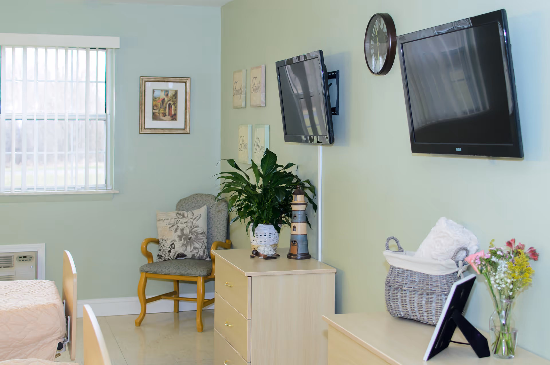 A cozy room with light green walls featuring two wall-mounted flat-screen TVs, a round wall clock, a wooden chair with a floral cushion, a dresser with a potted plant and decorative lighthouse, and a basket with a white blanket. There is a window with vertical blinds allowing natural light into the room.