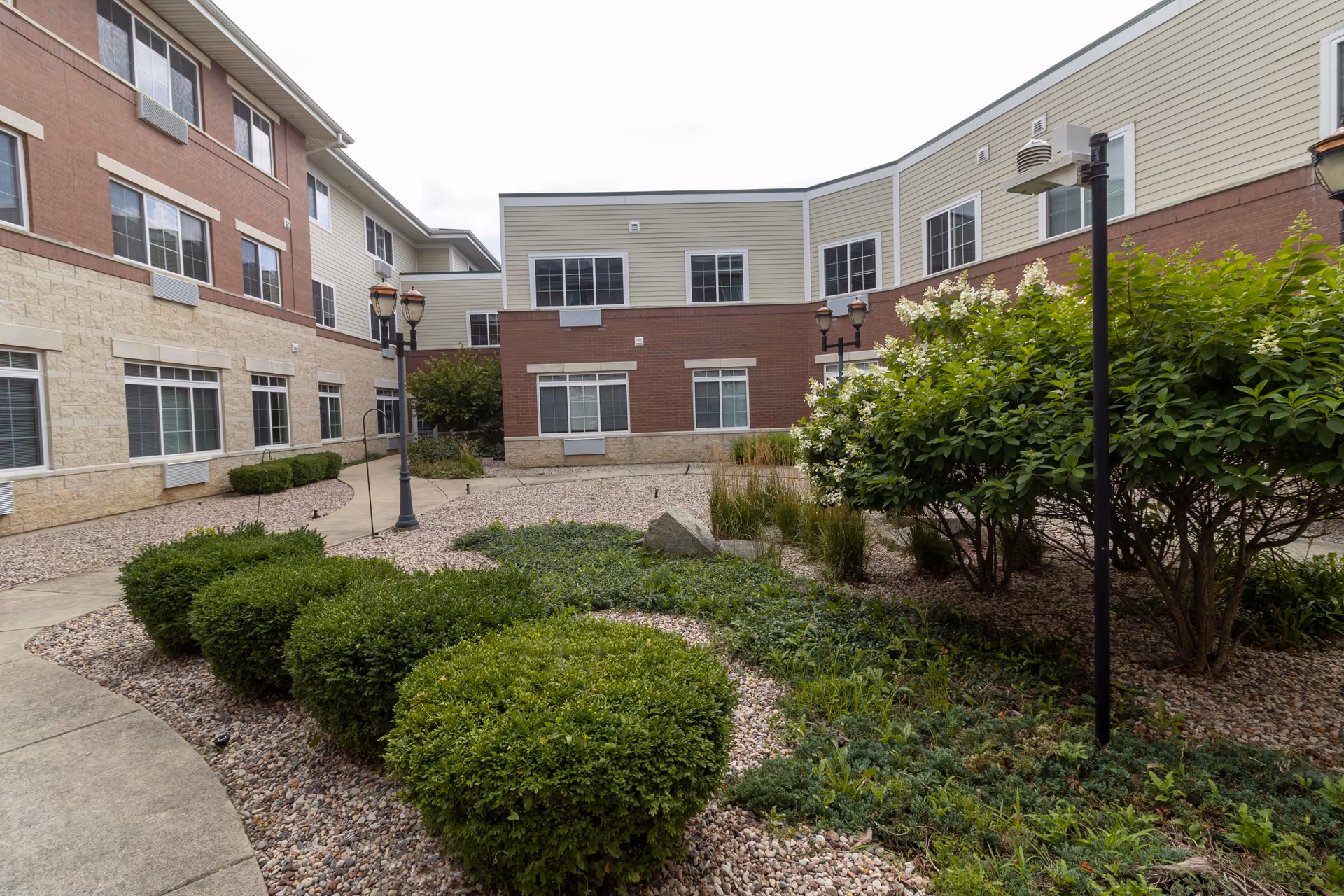 Outdoor courtyard area of a senior living facility with neatly trimmed bushes, flowering shrubs, and lamp posts surrounded by a three-story building with multiple windows.