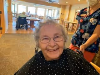 An elderly woman with glasses and curly gray hair smiling while seated in a wheelchair inside a well-lit room. A caregiver wearing a floral top and red pants stands behind her, pushing the wheelchair. In the background, there are tables and chairs arranged in a spacious common area with large windows letting in natural light.