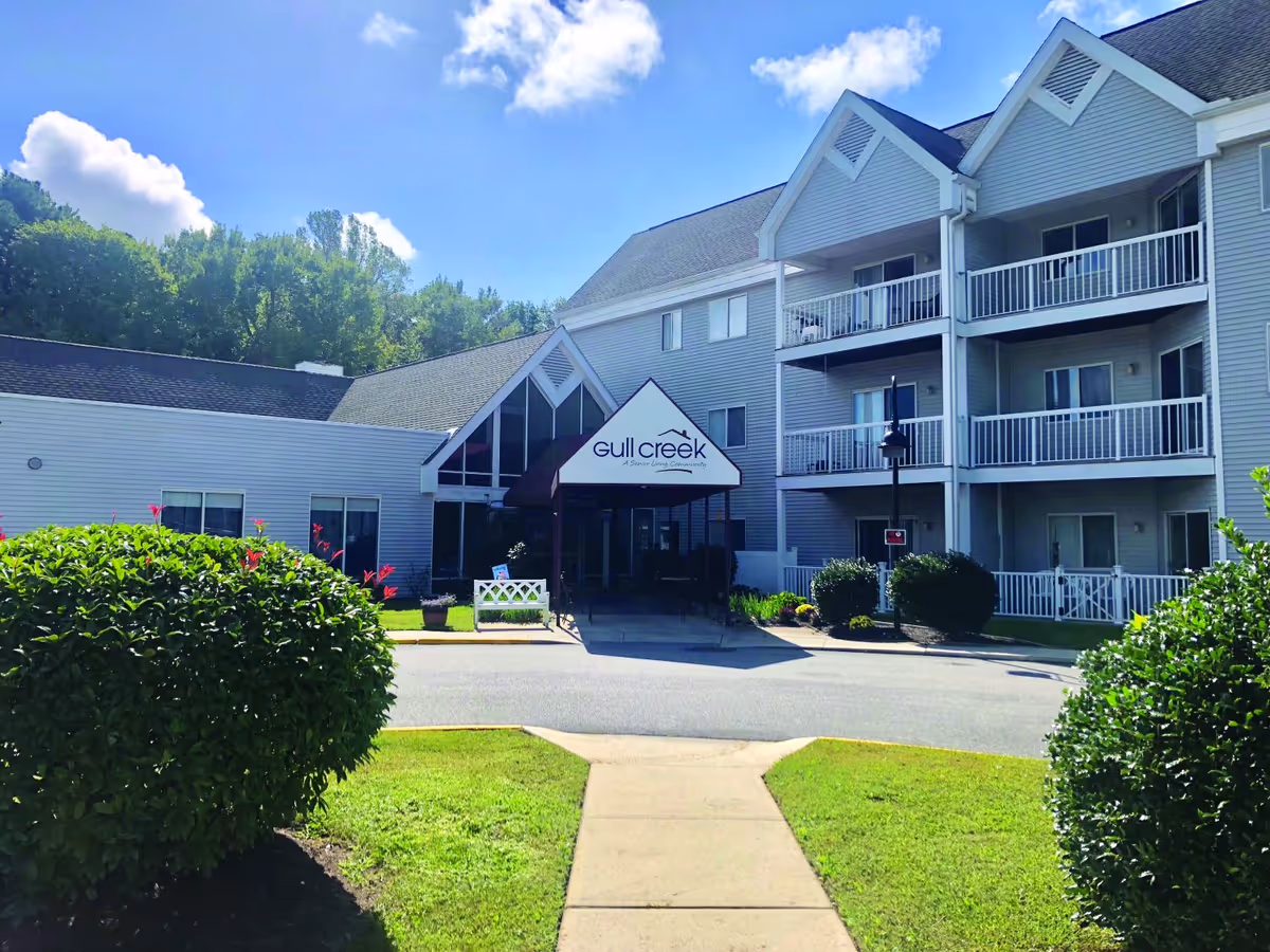 Exterior view of Gull Creek senior living community building with a covered entrance, balconies, and well-maintained landscaping including bushes and a lawn under a clear blue sky.