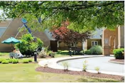 Courtyard outside a brick senior living building with trees, patio seating and umbrellas along a curved driveway and walkway.