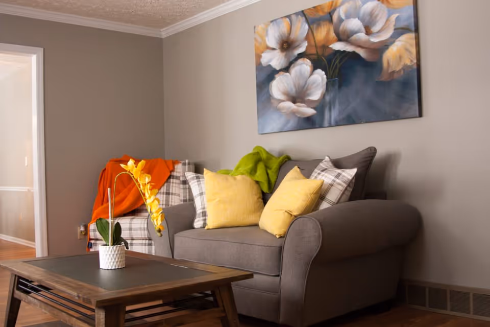 A cozy living room with a gray armchair and a plaid armchair, both adorned with yellow and plaid pillows and colorful throws. A wooden coffee table with a small white pot holding a yellow orchid is in front of the chairs. A large floral painting hangs on the beige wall above the gray armchair.