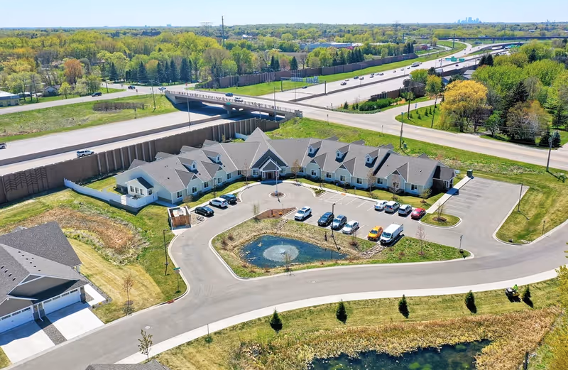 Aerial view of a senior living facility named Suite Living Memory Care & Assisted Living, showing a large building with multiple peaked roofs, a circular driveway with parked cars, a small pond with a fountain in the center, and surrounding green lawns. In the background, there is a highway with vehicles and a distant city skyline.