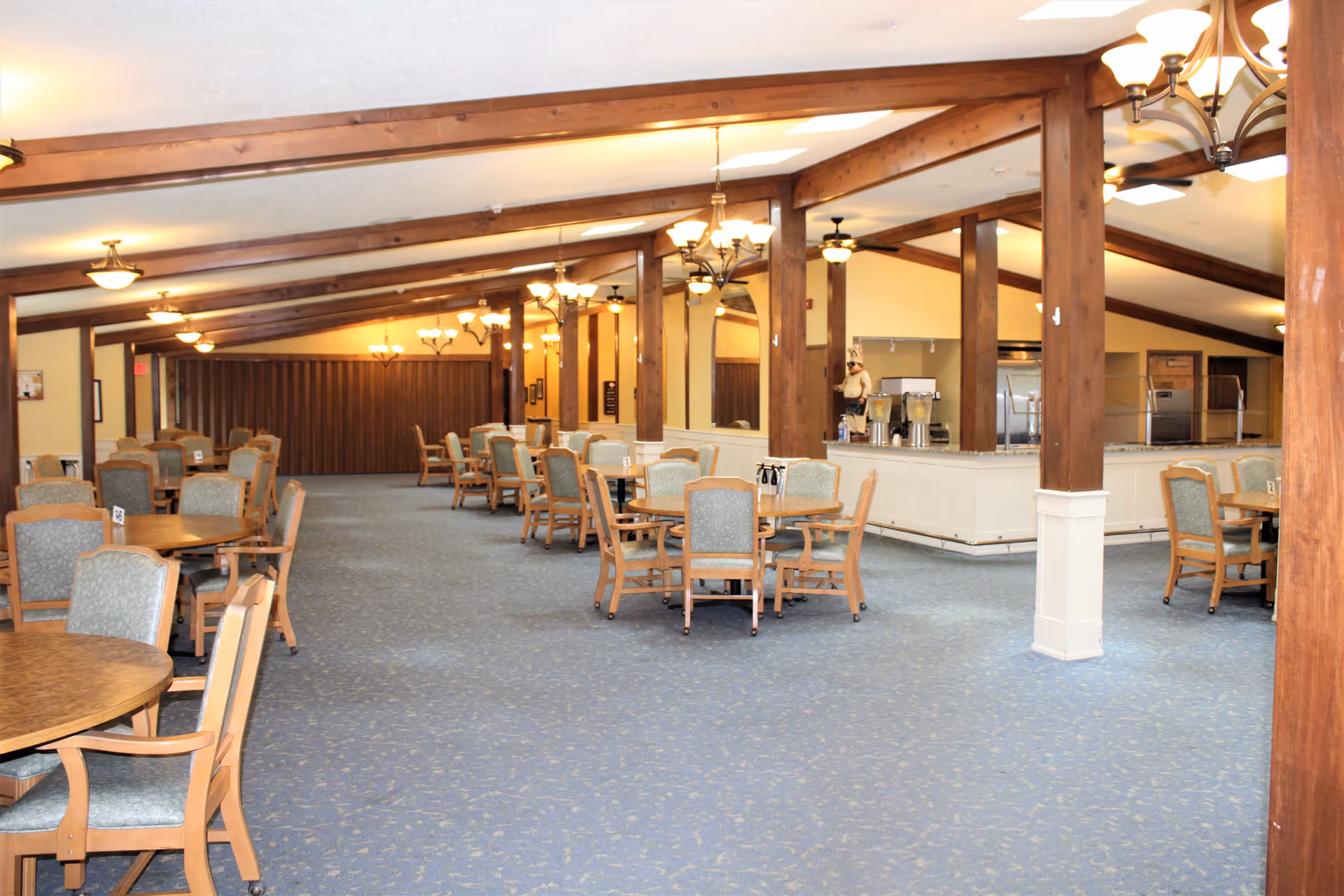 A spacious dining room with multiple round wooden tables and cushioned chairs arranged neatly. The room features wooden beams on the ceiling, several chandeliers, and a carpeted floor. In the background, there is a counter area with a person standing behind it, possibly serving or preparing food.