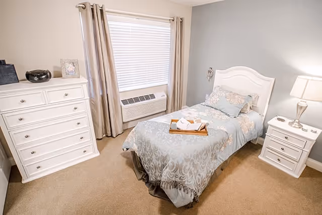 Comfortable single bedroom with a twin bed dressed in blue floral bedding, white dresser and nightstand, and a window air conditioner.