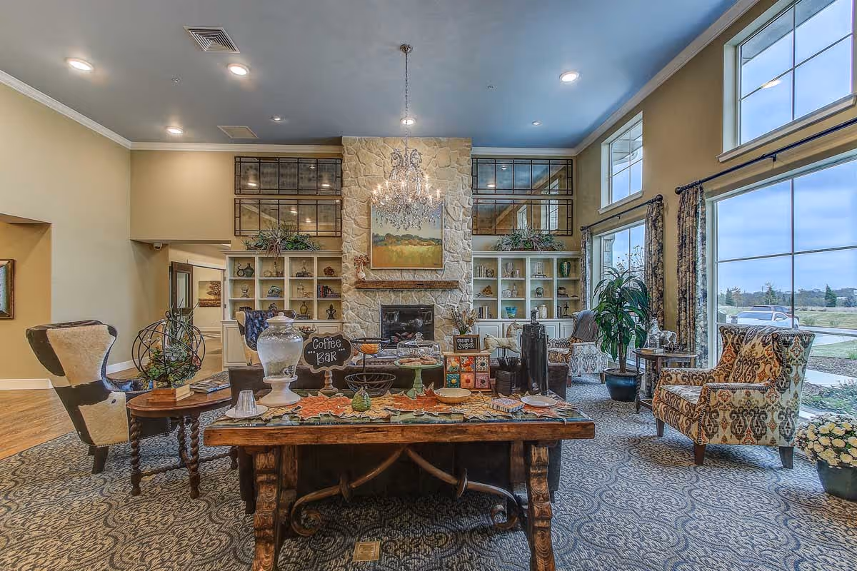 A spacious and well-lit living room area with large windows allowing natural light to fill the space. The room features a stone fireplace centered on the far wall, flanked by built-in shelves decorated with plants and ornaments. In front of the fireplace, there is a wooden table set up as a coffee bar with various items including a large glass jar, cups, and a small chalkboard sign. Comfortable upholstered chairs and armchairs are arranged around the room, with patterned carpeting covering the floor and a chandelier hanging from the ceiling.